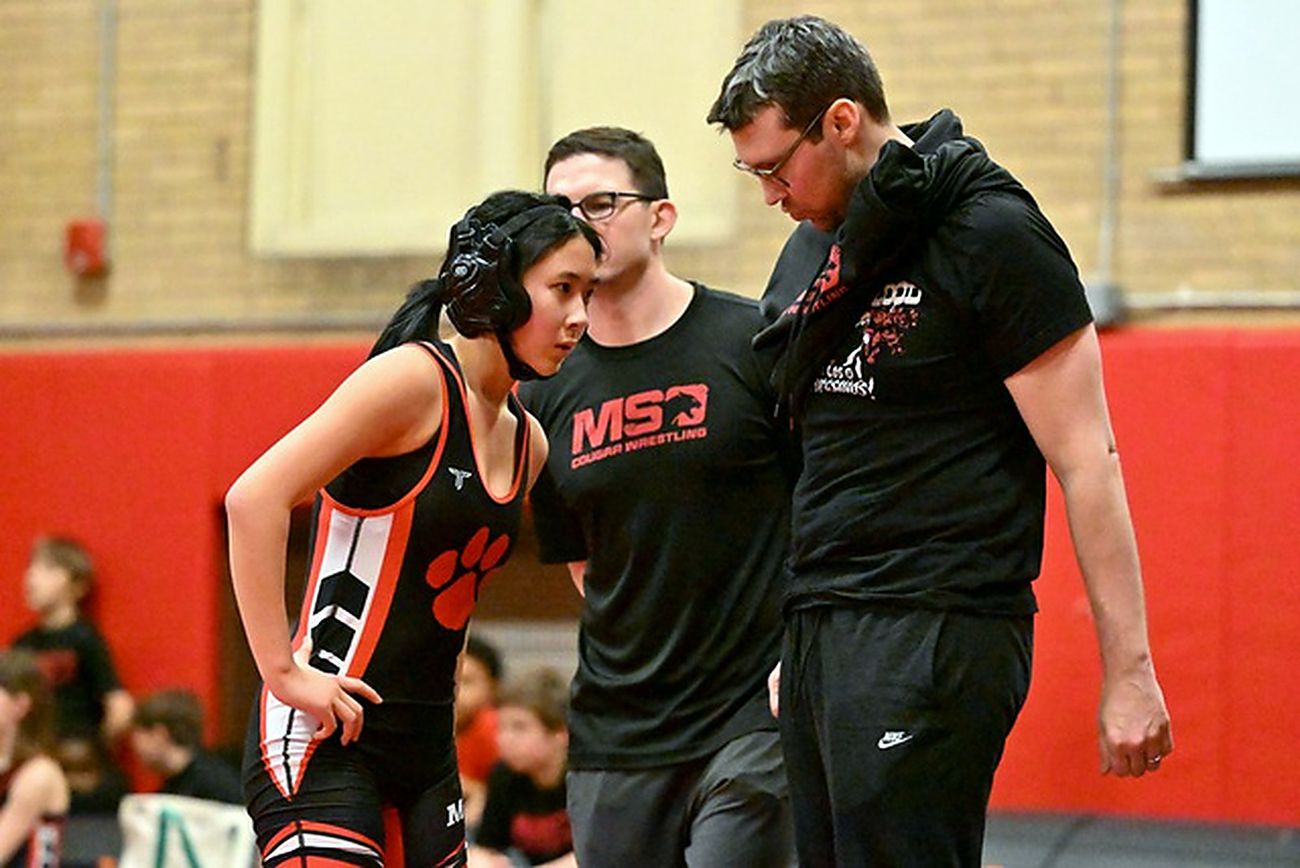 Two MSO coaches talking strategy with a girl wrestler at the edge of the mat.